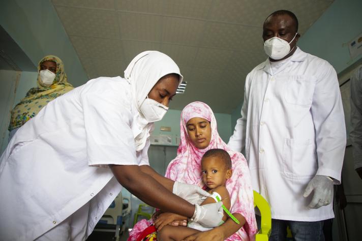 A woman on a chair holding her child. A nurse is measuring the arm of the child while a doctor stands in the back.