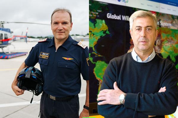Man on the left in pilot's uniform in front of a helicopter and man on the right standing in front of large screens with maps